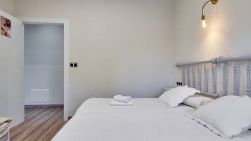 A bedroom with a bed, white bedding, a nightstand, a wall-mounted light, and a door leading to another room.