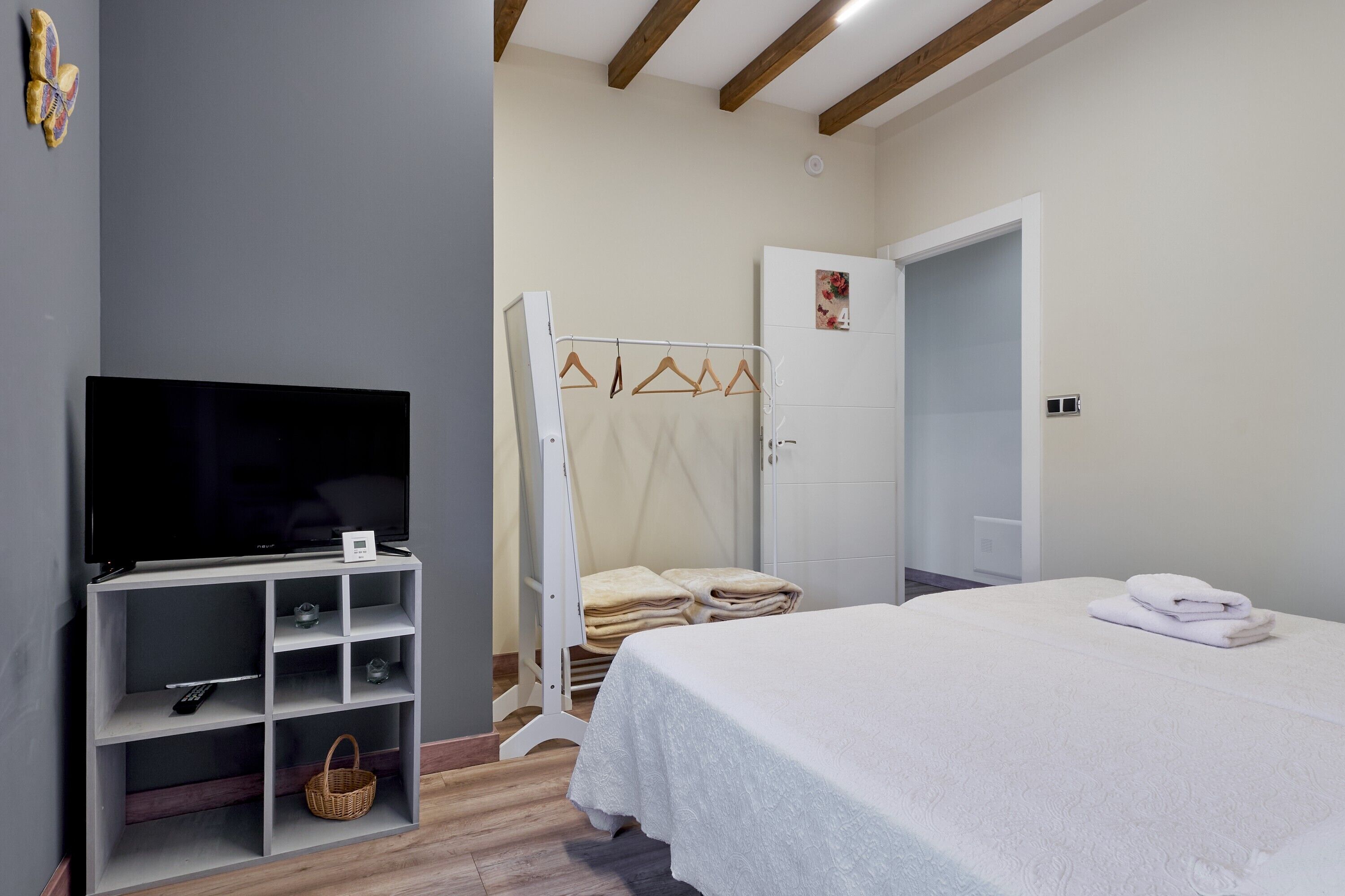Un dormitorio con televisor sobre un mueble, cama con colcha blanca, armario y puerta que da a otra habitación.