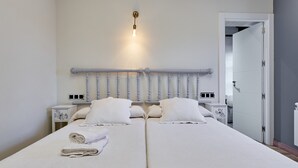 Un lit double avec du linge de lit blanc, une tête de lit dotée d’un porte-serviettes, des tables de chevet et une porte donnant sur une autre pièce.