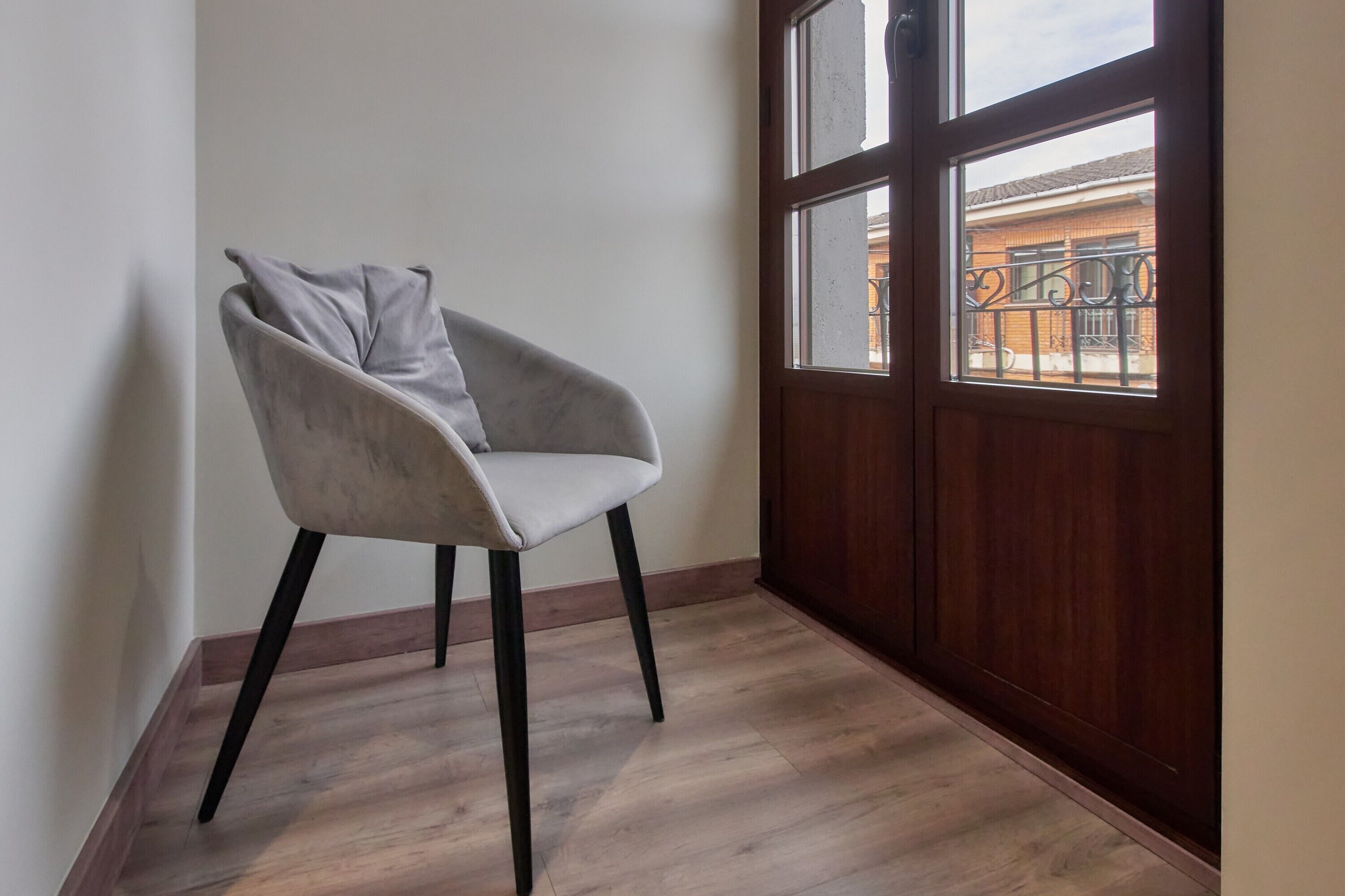Habitación de esquina con una silla, piso de madera, ventana con vista a un edificio y puerta de madera.