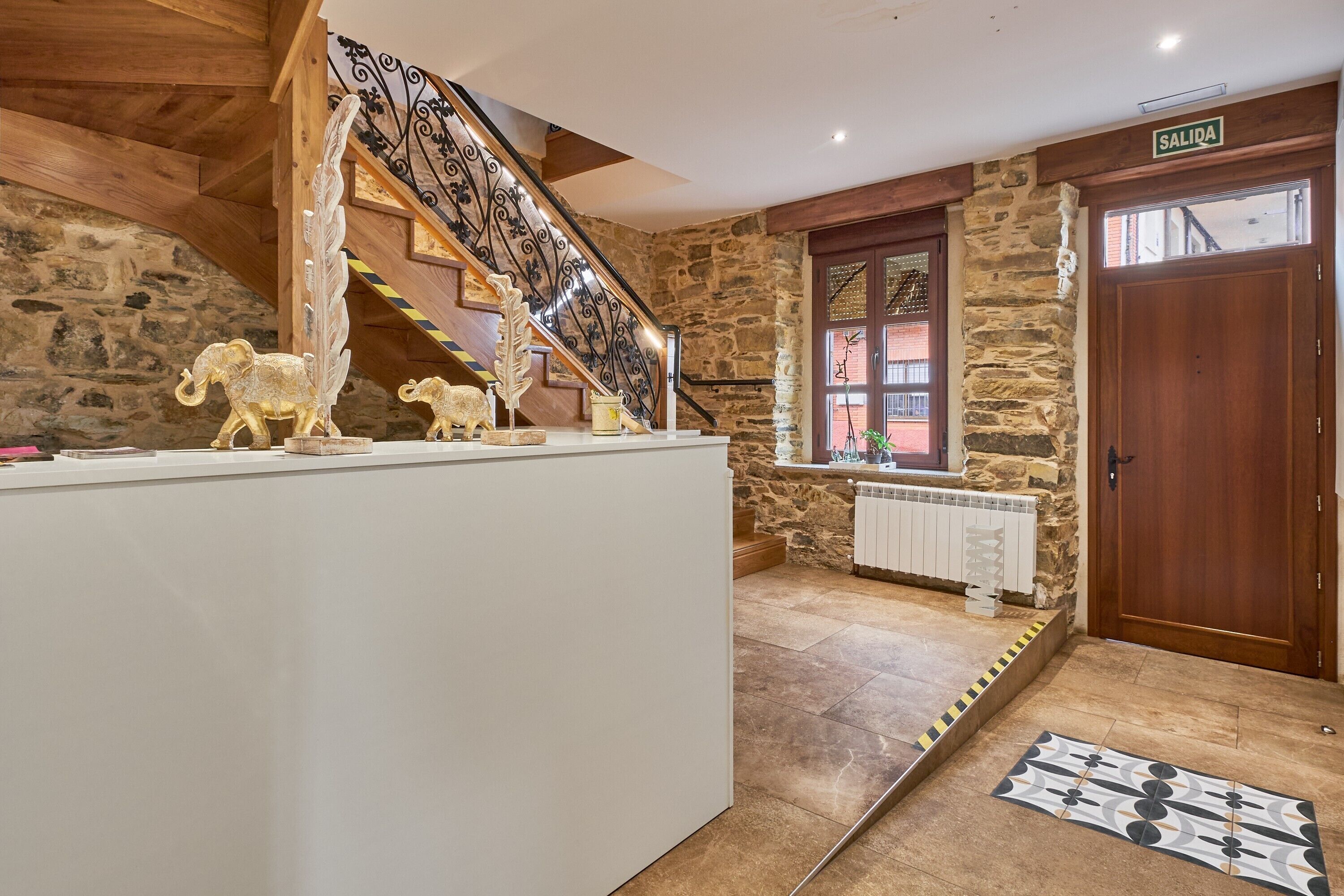 Un espace d’accueil avec un escalier, un mur en pierre, une porte en bois portant l’inscription « SORTIE » et un tapis à motifs sur le sol.