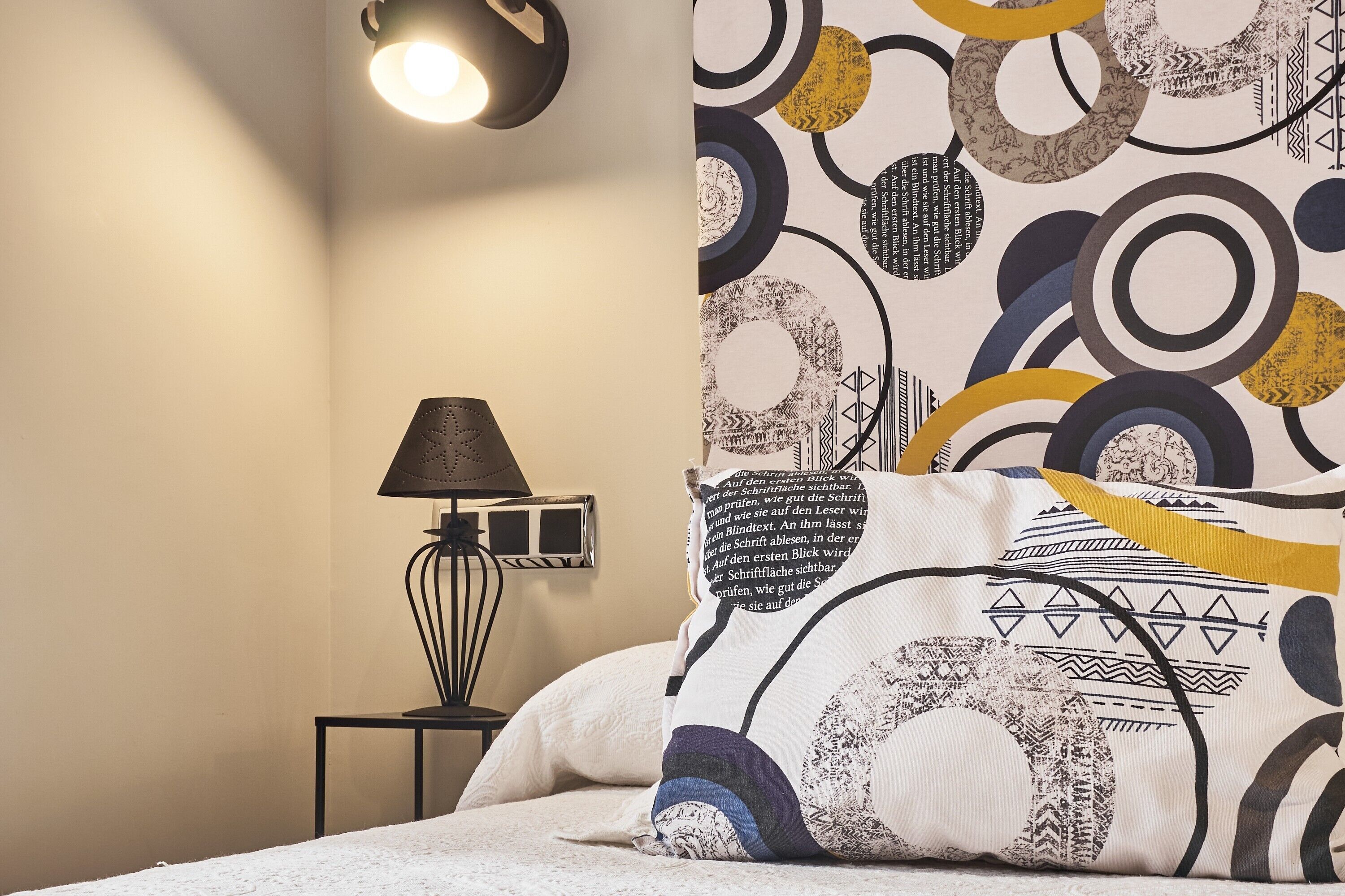 A bedroom with a bed, a nightstand with a lamp, and a wall with abstract patterns.