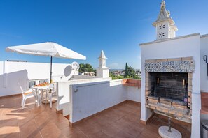 Eine Dachterrasse mit einem weißen Tisch, Stühlen, einem weißen Sonnenschirm, einem Kamin aus Ziegelsteinen und einem Glockenturm.