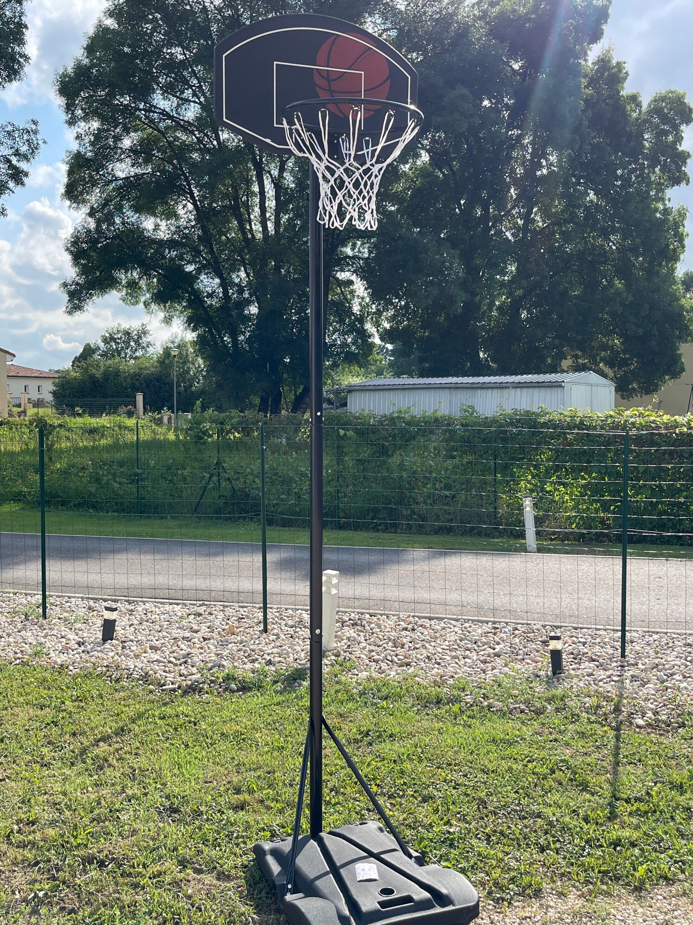 Een basketbalring in een open omgeving, met een hek, bomen en een gebouw op de achtergrond.