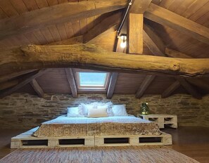 Una camera da letto rustica in mansarda, con soffitto spiovente, un lucernario, un letto di legno con biancheria a motivi decorativi e una parete in pietra.