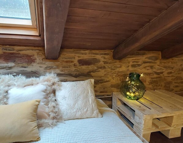 Una camera accogliente con parete in pietra, soffitto in legno, una finestra, un letto con coperta bianca, un cuscino e un piccolo tavolo di legno con una bottiglia verde.