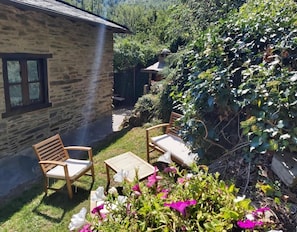 A stone building with a chimney, a small patio with two wooden chairs and a table, and a garden with various plants and flowers.