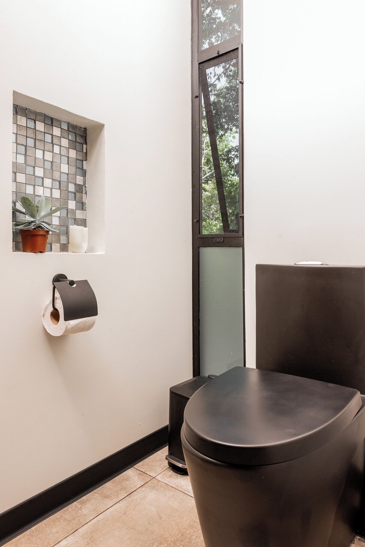 A small bathroom with a toilet, a trash bin, a toilet paper holder, and a window with a view of trees.