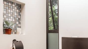 A small bathroom with a toilet, a trash bin, a toilet paper holder, and a window with a view of trees.