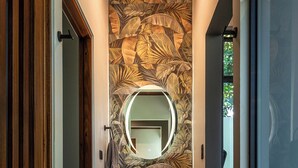 Un pasillo estrecho con una puerta de madera, un espejo redondo, un lavamanos negro y un estante con toallas y una planta en maceta.
