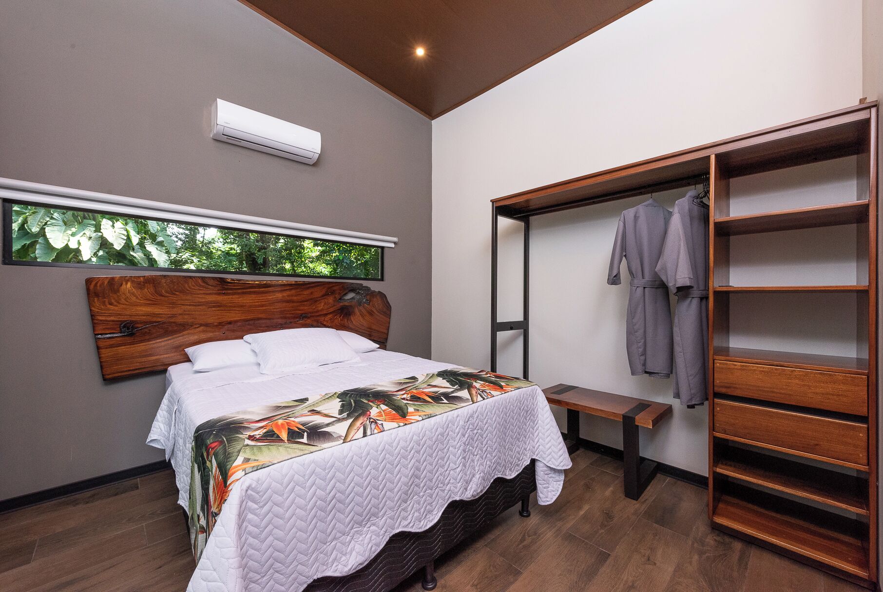 A bedroom with a bed, a wooden headboard, a large window with a view of trees, a built-in bench, and a wooden wardrobe with hanging clothes.