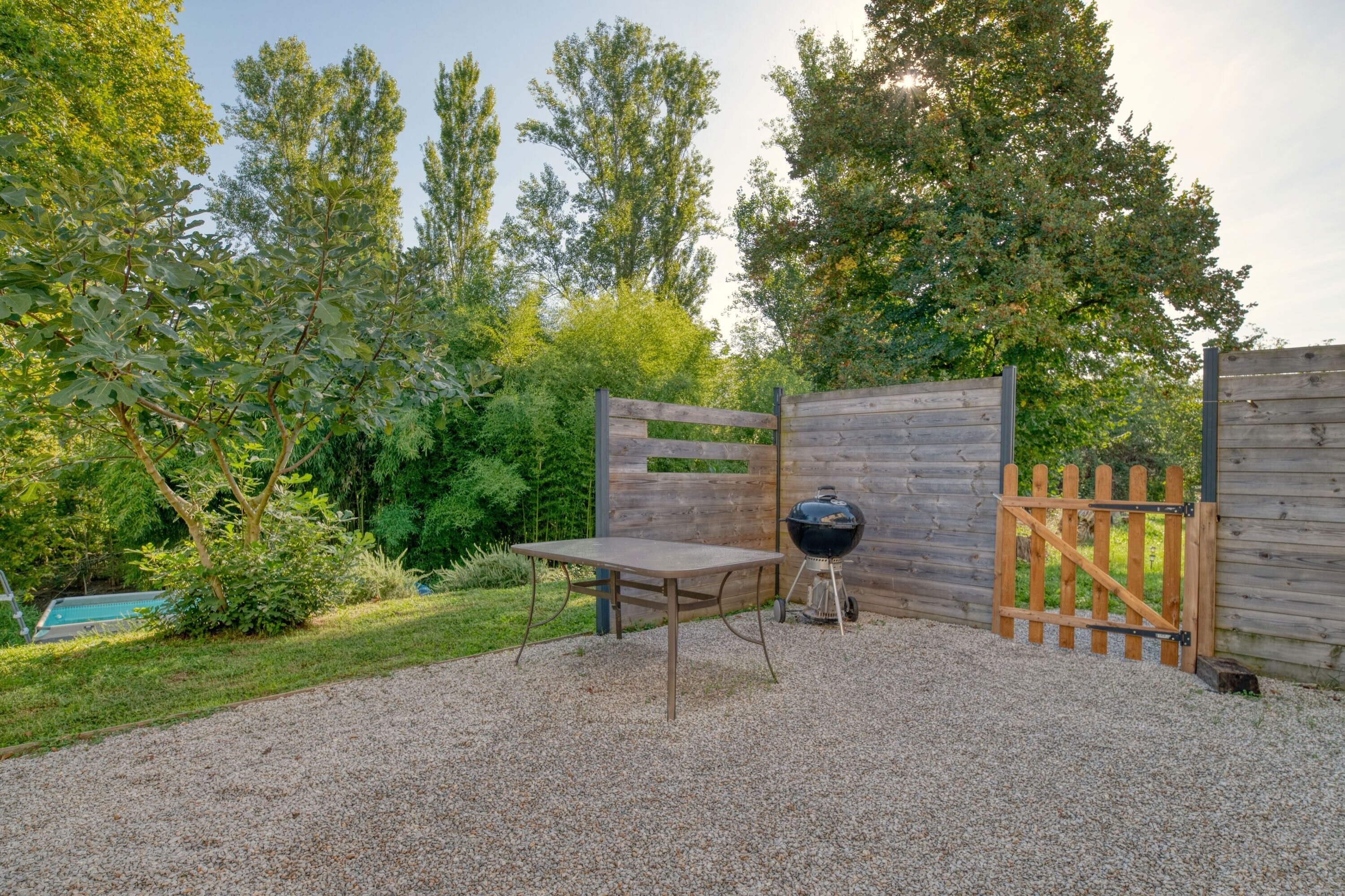 Eine gepflegte Schotterterrasse mit Holztisch und Grill, umgeben von einem Holzzaun und üppigen Bäumen.