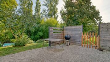 Une terrasse en gravier avec une table en bois et un barbecue, entourée d’une clôture en bois et d’arbres touffus.