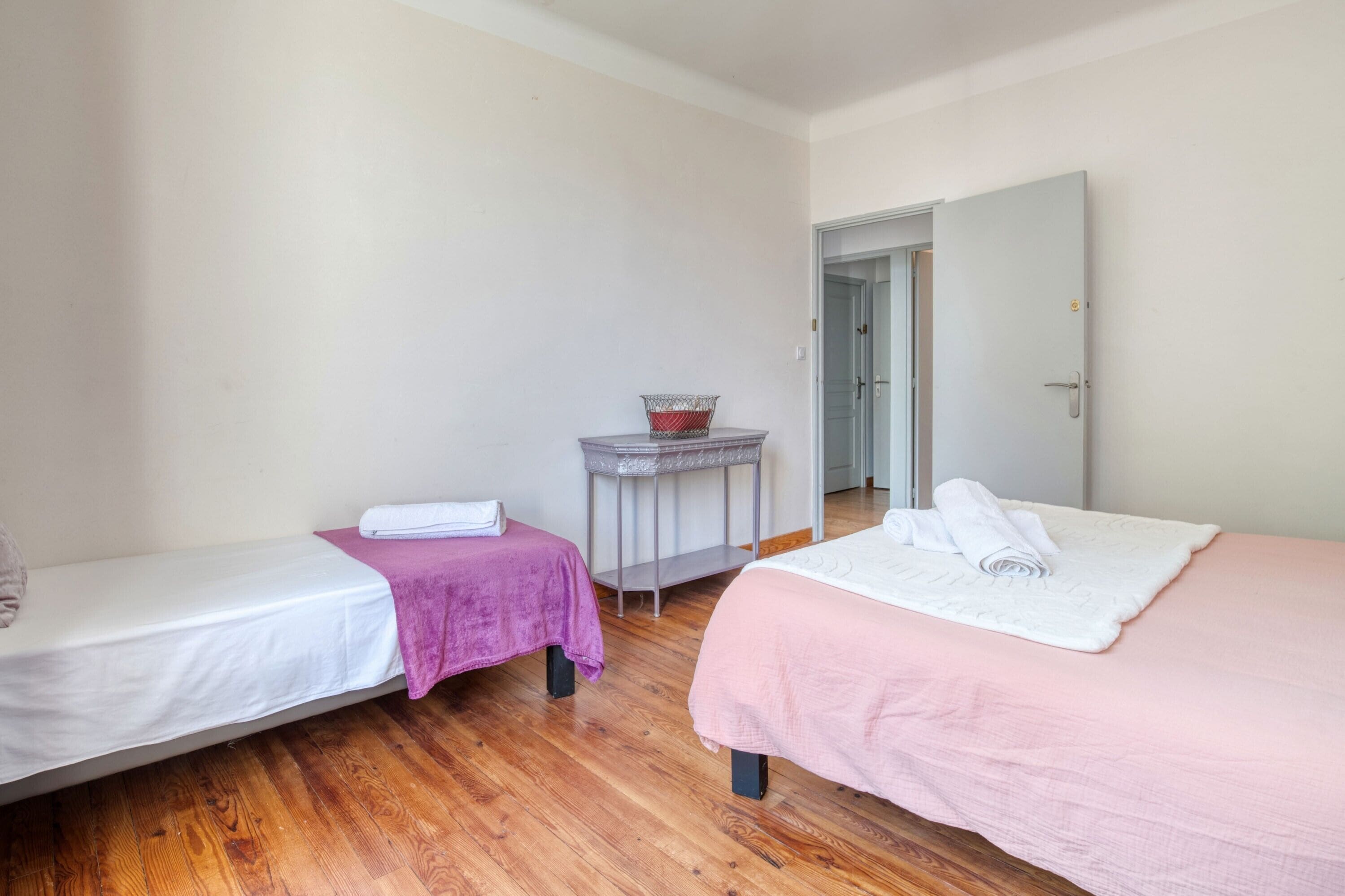 Une chambre avec deux lits simples, une petite table avec un panier, et une porte donnant sur une autre pièce.