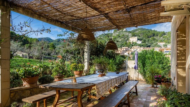 Eine Terrasse im Freien mit Tisch und Bänken, umgeben von Blumentöpfen und Blick auf eine Hügellandschaft.