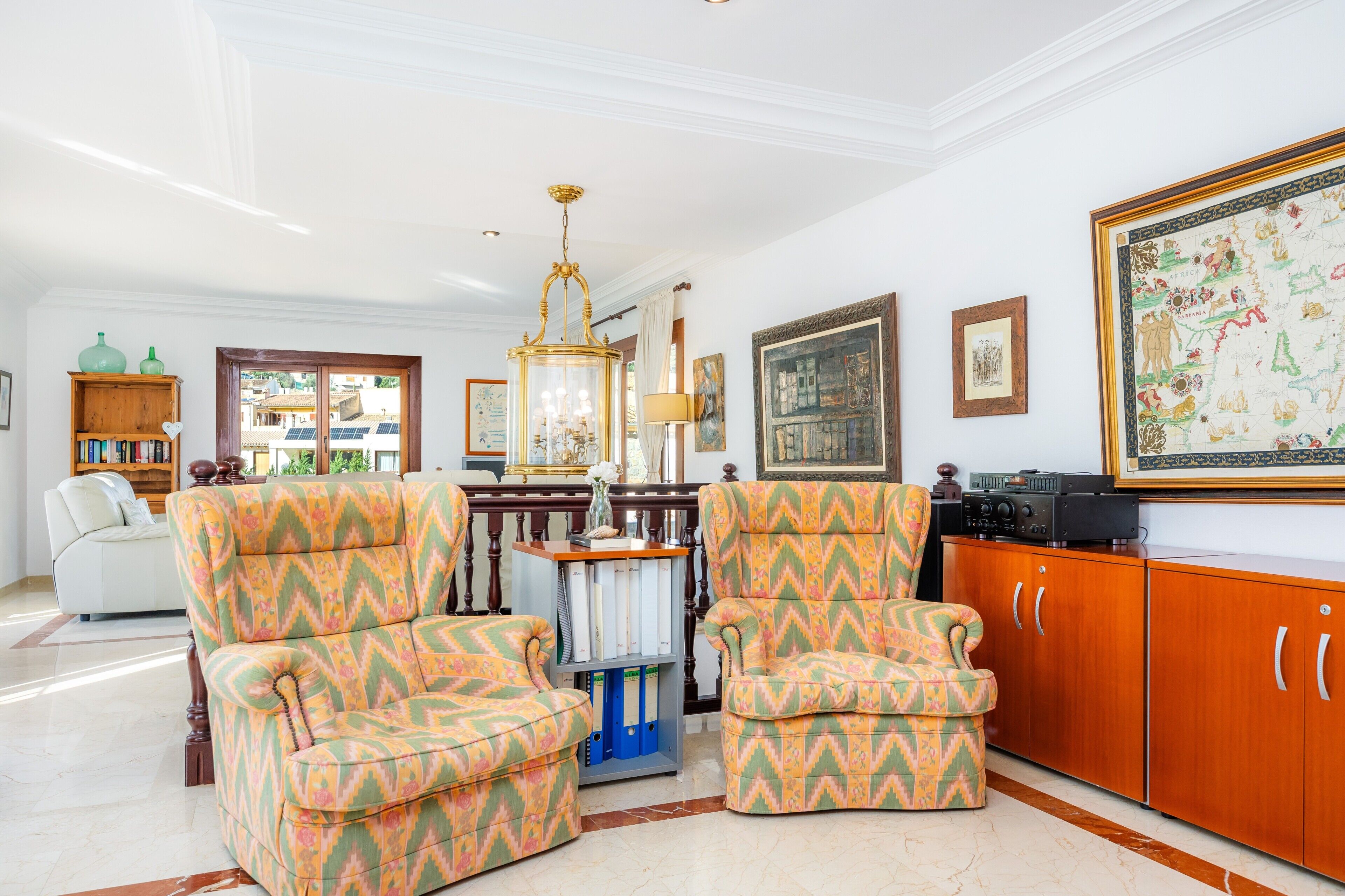 A living room with patterned armchairs, a wooden cabinet, a chandelier, and framed artwork.