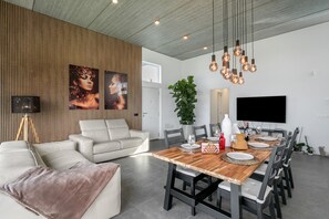 Un espace de repas moderne avec une table en bois, des chaises noires, un canapé blanc et un grand téléviseur fixé au mur.