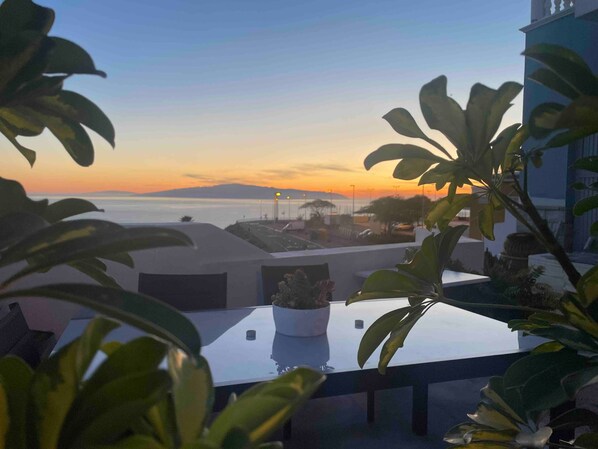 Una terraza con vistas a la puesta de sol sobre el mar, plantas en macetas y un edificio al fondo.