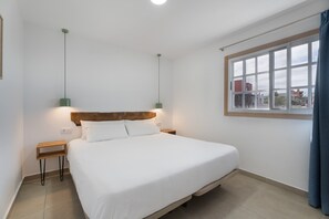 Una cama individual con cabecera de madera, dos mesitas de noche con lámparas, una ventana con vistas a edificios y una cortina a la derecha.