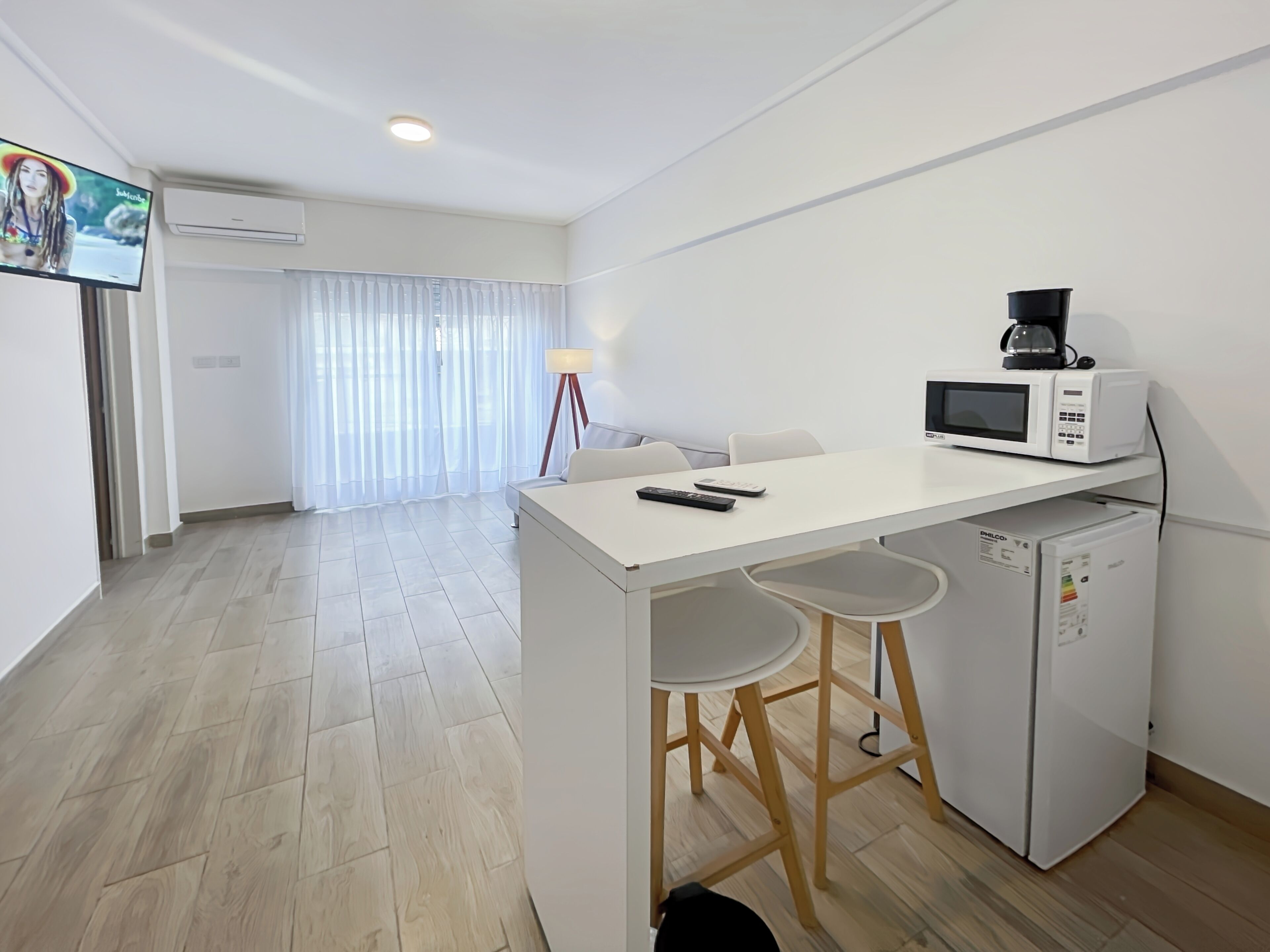 Une cuisine moderne avec un plan de travail blanc, un four à micro-ondes, une machine à café et un réfrigérateur. On y trouve deux tabourets de bar et une petite table.