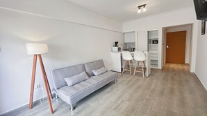 Una sala moderna con un sofá gris, una lámpara de pie y una zona de cocina con microondas y taburetes.