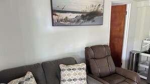 Uma sala de estar com um sofá cinzento, almofadas estampadas, uma poltrona marrom e um quadro pendurado na parede.