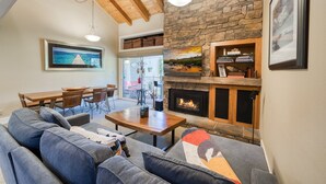 A cozy living room with a stone fireplace, wooden ceiling beams, and a dining area with a table and chairs.