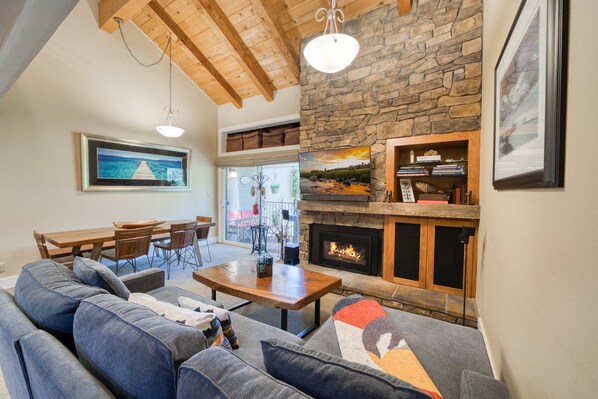 A cozy living room with a stone fireplace, wooden ceiling beams, and a dining area with a table and chairs.