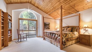 A bedroom with a four-poster bed, a wooden ceiling, a large window, a bookshelf, and a nightstand with a lamp.
