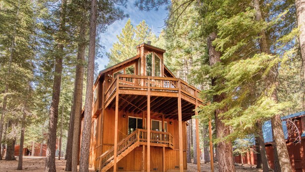 A wooden cabin with a balcony and stairs, surrounded by tall pine trees.