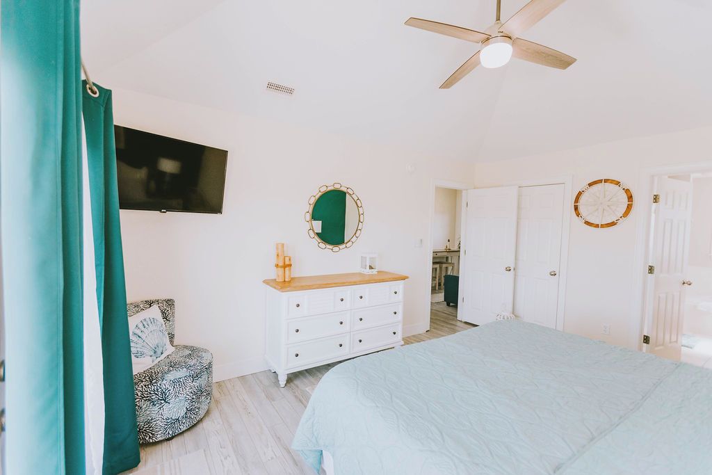 A bedroom with a bed, a dresser, a mirror, a ceiling fan, and a TV on the wall.