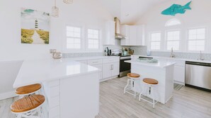 A modern kitchen with white cabinetry, a central island with stools, and a large window. - Coastal Distancing: Oceanside, Private Pool, Hot Tub, Close to Beach (Nags Head)