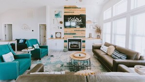 A spacious living room with teal and brown sofas, a fireplace, and a large TV mounted on a wooden wall. - Coastal Distancing: Oceanside, Private Pool, Hot Tub, Close to Beach (Nags Head)