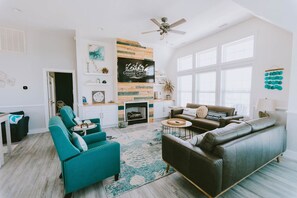 A spacious living room with a fireplace, teal and brown sofas, and a large window. - Coastal Distancing: Oceanside, Private Pool, Hot Tub, Close to Beach (Nags Head)