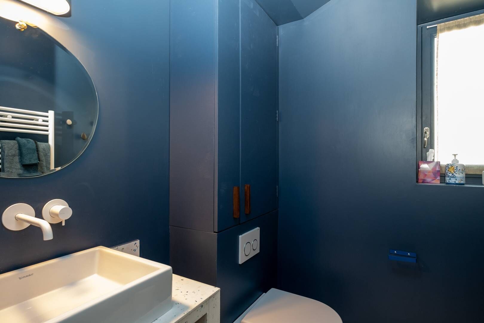 A bathroom with a white sink, a mirror, and a toilet. There is a window with a view of the outside.