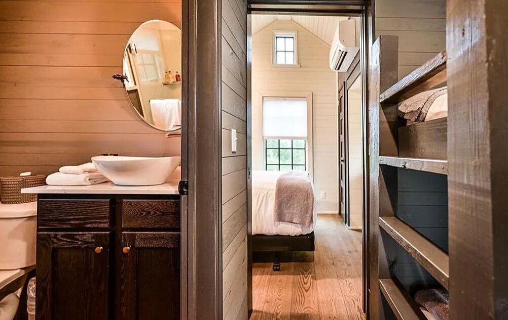 A narrow hallway leading to a bedroom with a single bed, a small window, and a wooden floor.