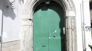 A green door with a number plate, set within a stone archway, attached to a white wall with a brick base.