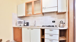 A compact kitchen with white cabinets, a built-in stove, a kettle, and a small refrigerator.