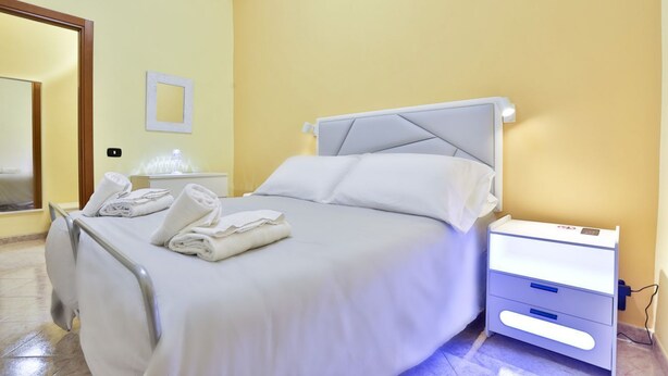 A neatly made bed with white linens, a bedside table with a lamp, and a small drawer unit.