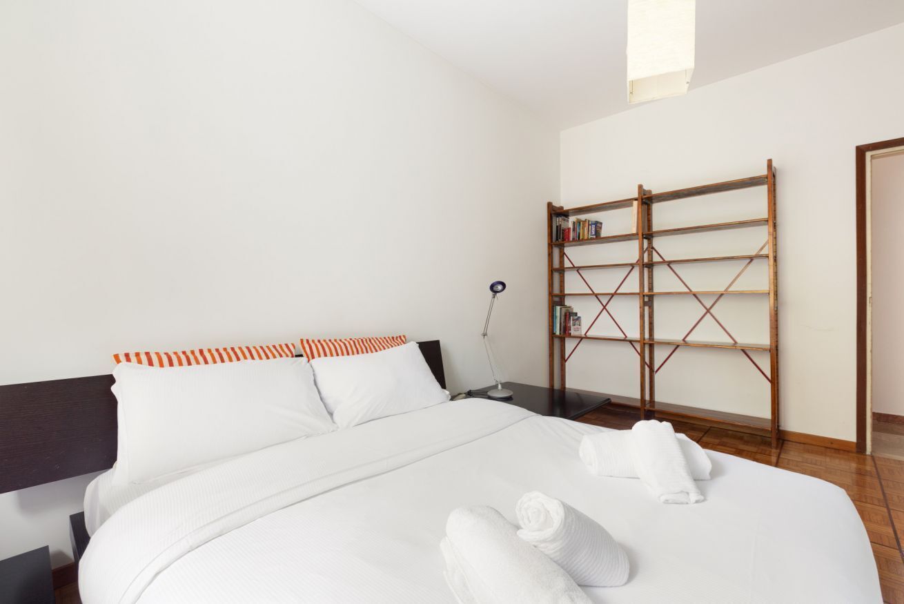 A neatly made bed with white linens and rolled towels, a wooden bookshelf with books, and a nightstand with a lamp.