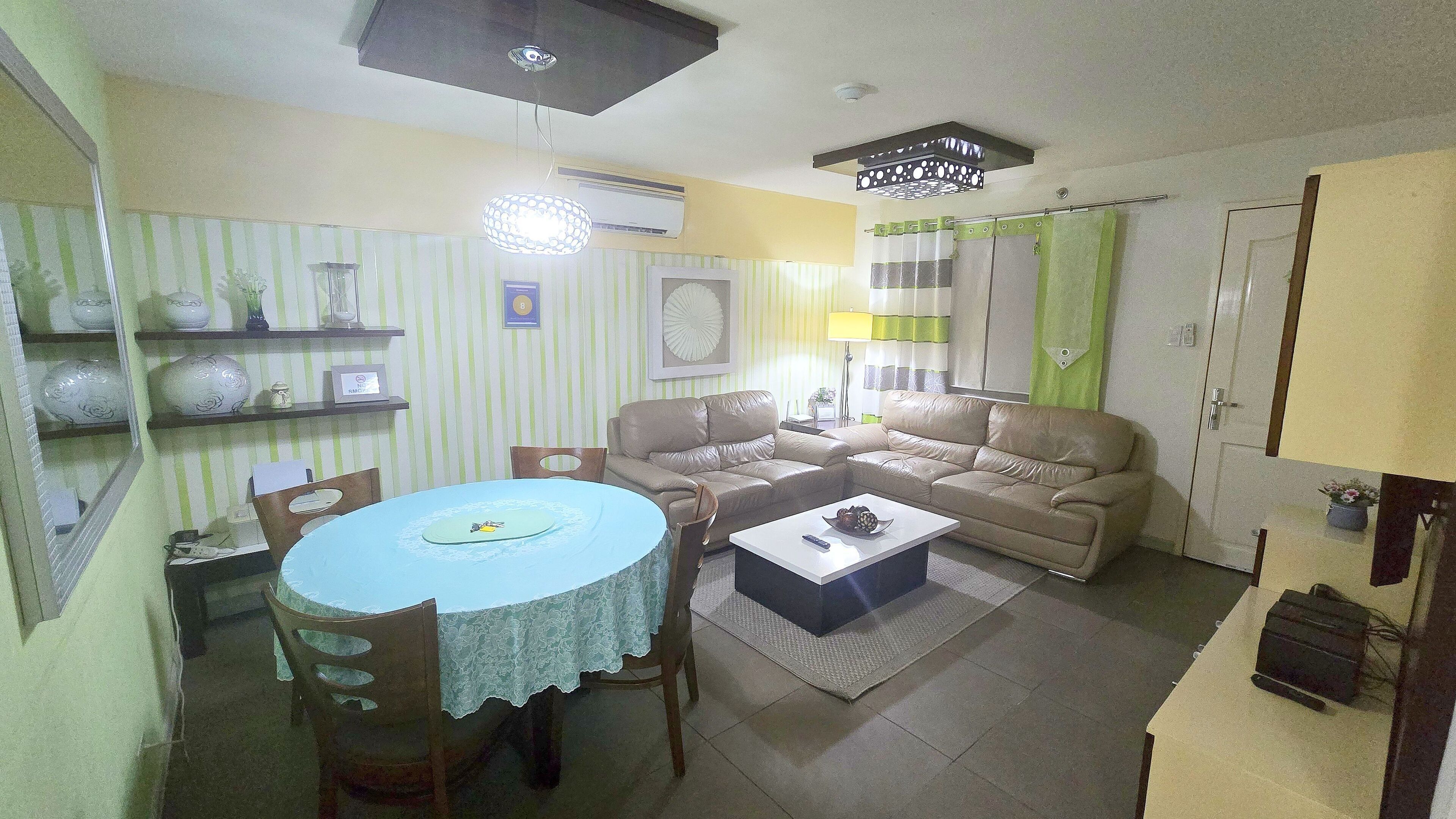 A living room with a sofa, a coffee table, and a dining table with chairs. There is a lamp, a wall-mounted air conditioner, and a door with a green curtain.