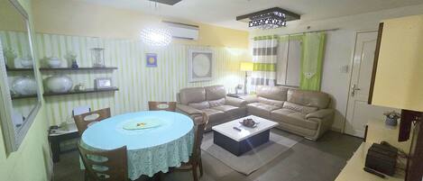 A living room with a sofa, a coffee table, and a dining table with chairs. There is a lamp, a wall-mounted air conditioner, and a door with a green curtain.