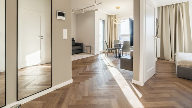 Una habitación de hotel moderna con suelo de madera tipo espiga, un sofá, una zona de comedor con mesa y sillas, y un espejo grande.