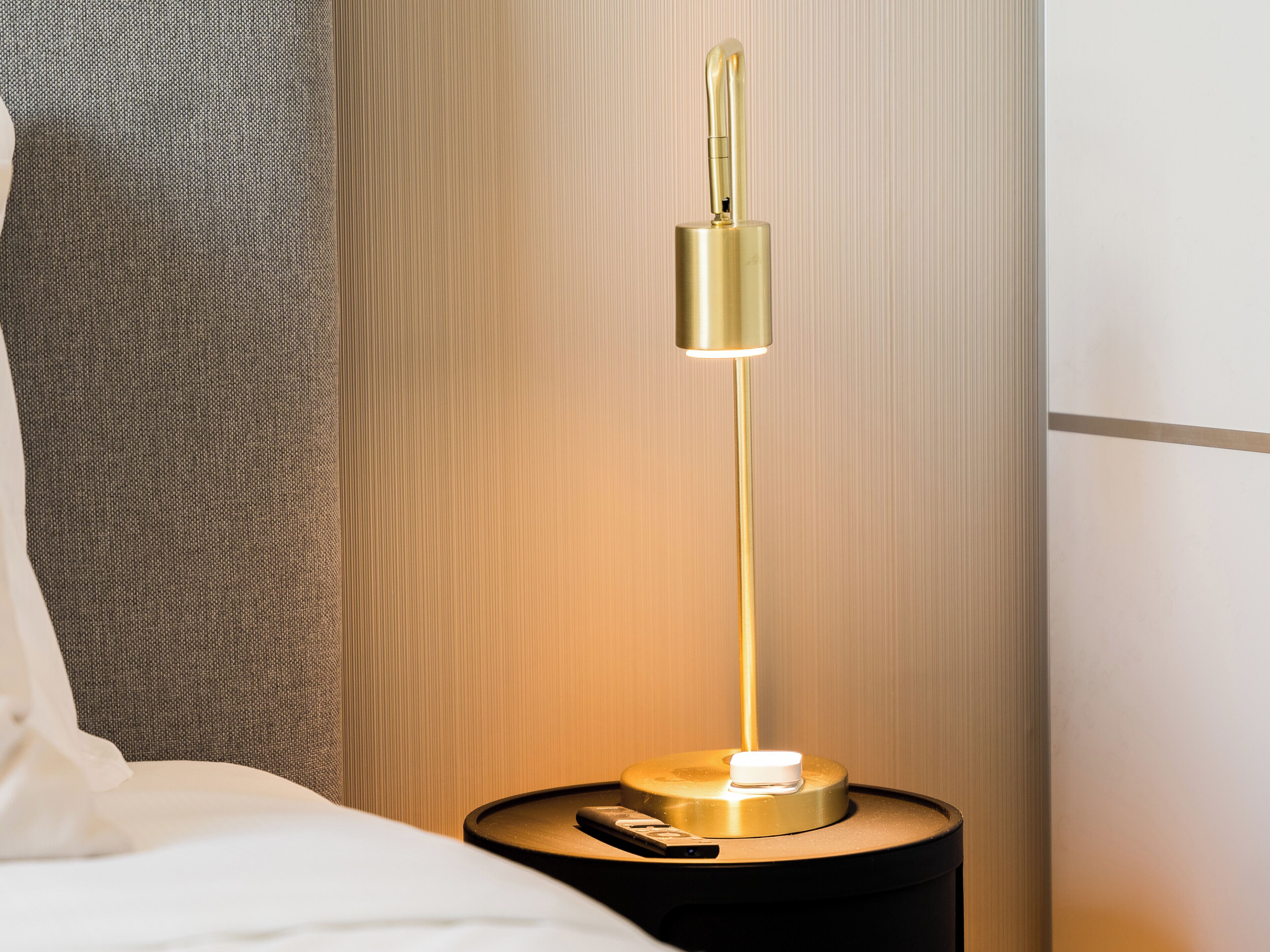 Une table de chevet avec une lampe de couleur dorée, un oreiller blanc et une petite table d’appoint noire.