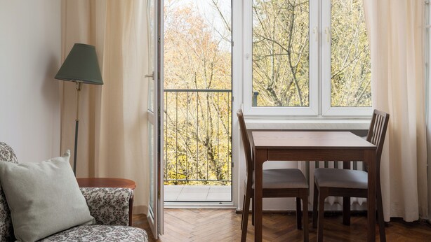 Een kamer met een eettafel en stoelen, een bank, een lamp en uitzicht op bomen door een raam.