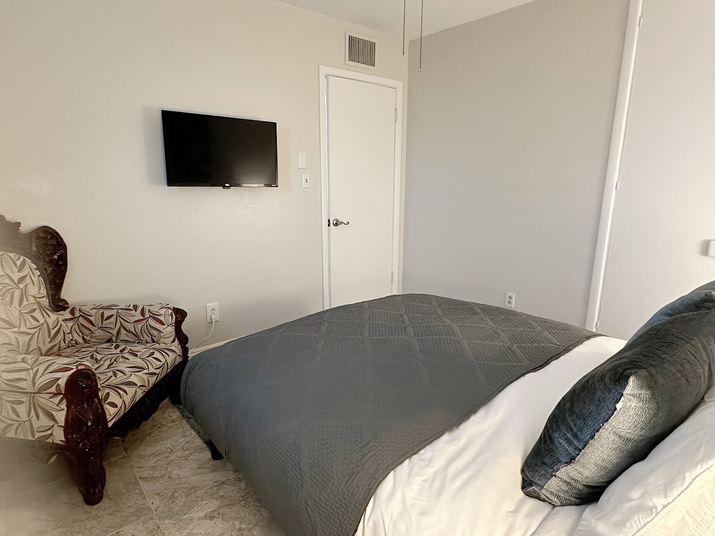 Ein Schlafzimmer mit einem Bett, einem Sessel, einem an der Wand befestigten Fernseher und einer Tür.