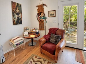 A living room with a leather armchair, a small round table with books and a lamp, a bookshelf, and a large window with a view of greenery.