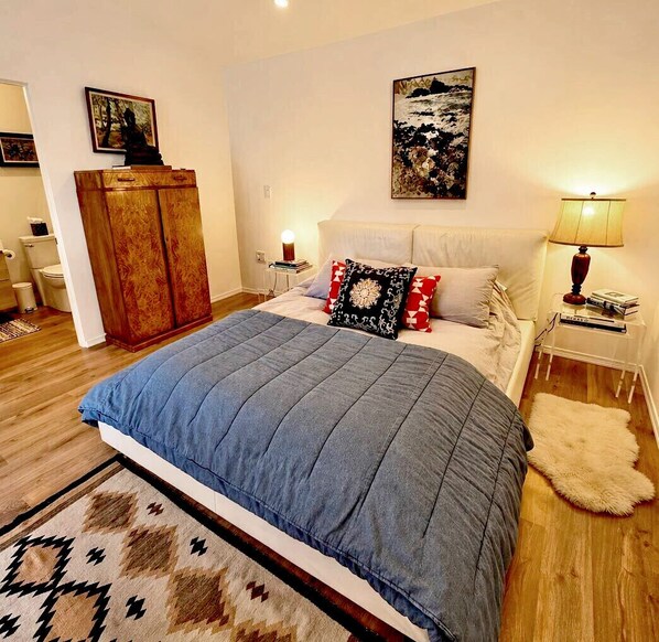 A bedroom with a bed, a nightstand, a lamp, a wardrobe, and a painting on the wall. - Private Garden Cottage • Walk to Downtown Paso (Paso Robles)