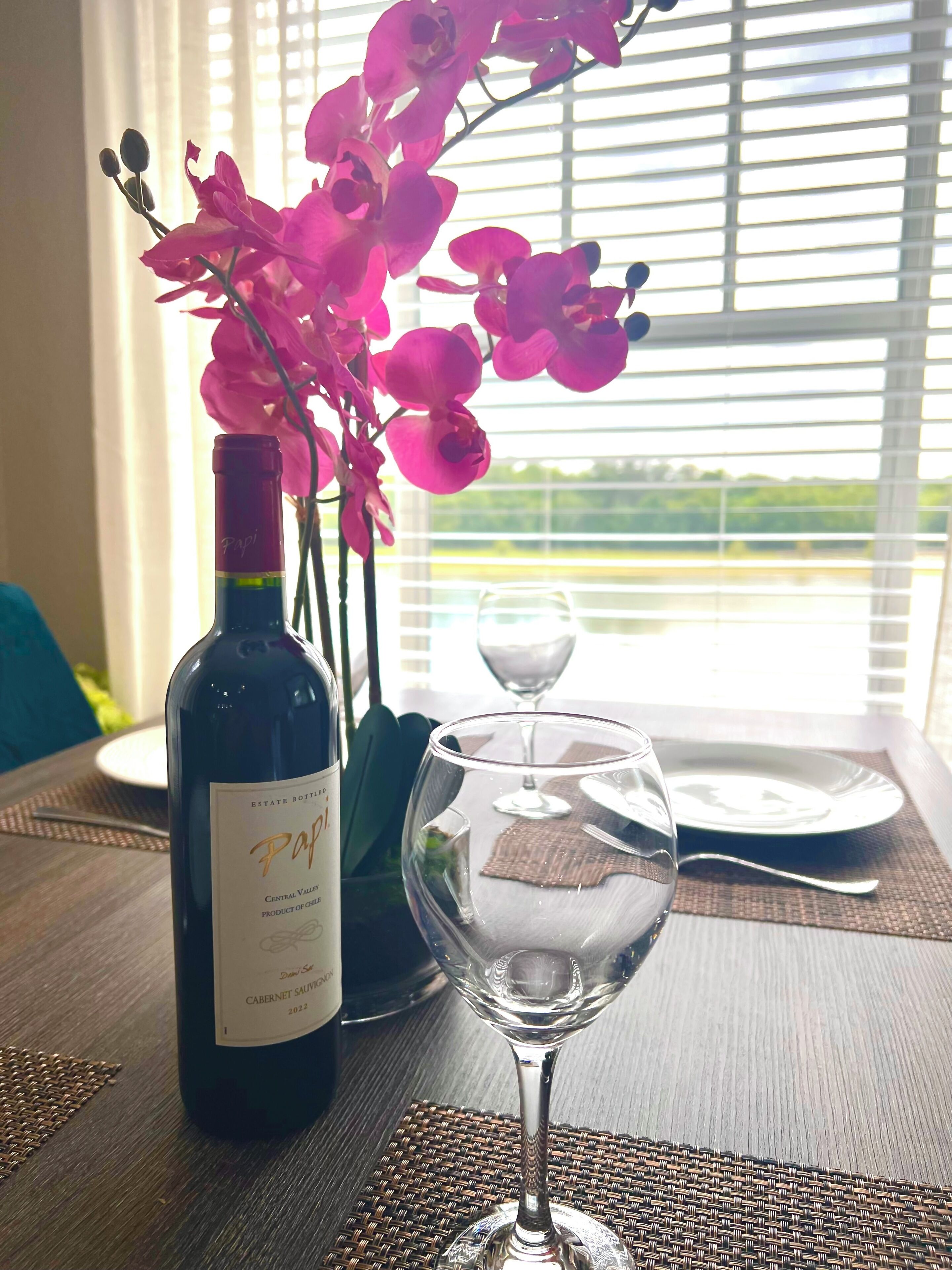 Une bouteille de vin, un verre et des orchidées roses sur une table, avec vue sur la verdure à travers les stores.