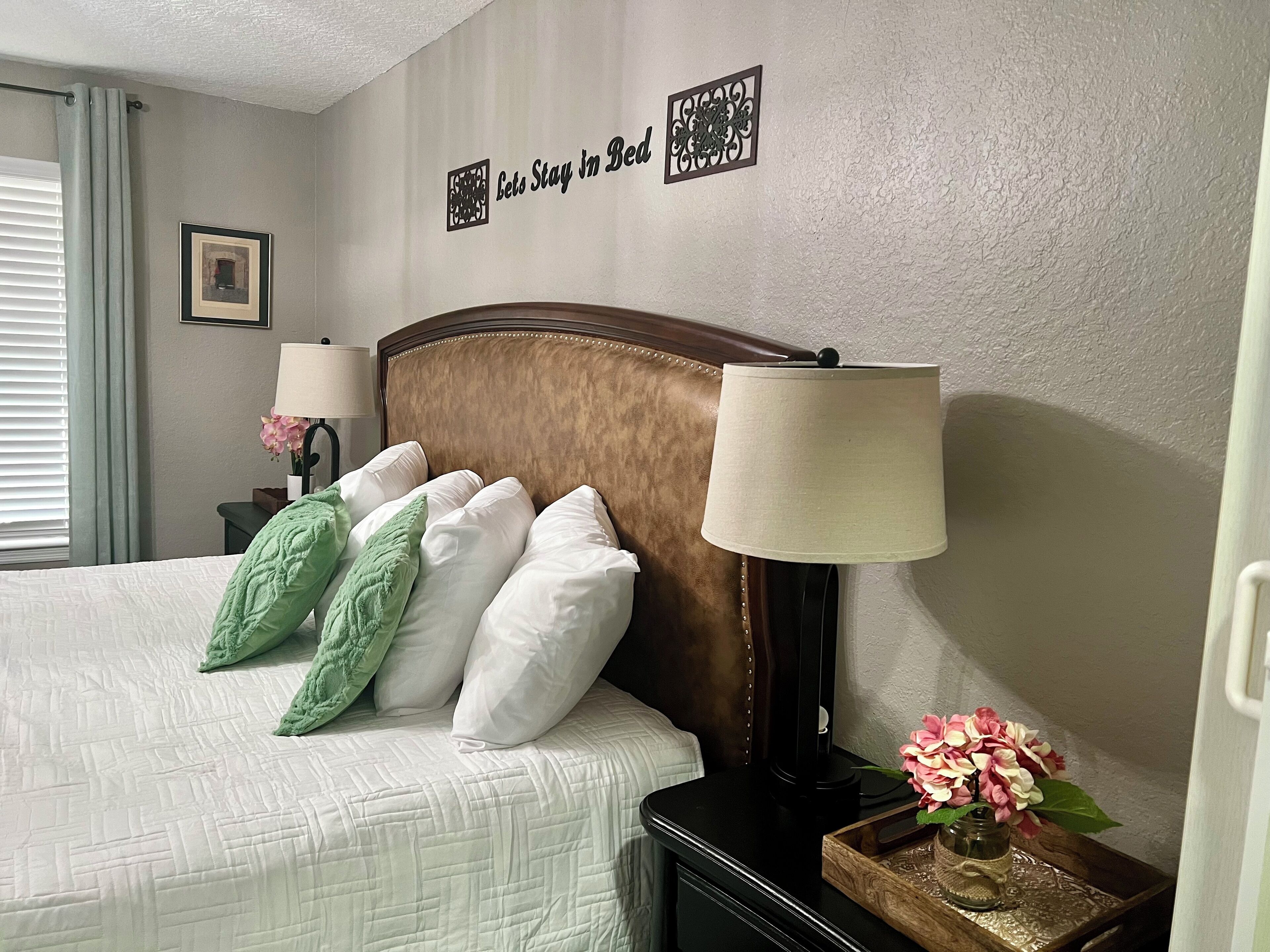 A neatly made bed with white and green pillows, a wooden headboard, a bedside lamp, and a small table with a vase of flowers.
